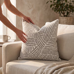 Cargar imagen en el visor de la Galería, Manos acomodando el cojín decorativo Celine Gray 50x50cm con diseño geométrico sobre un sillón beige.