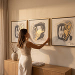 Cargar imagen en el visor de la Galería, Mujer colocando el set de 3 cuadros decorativos Pandora con hoja de oro en la pared.