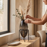 Cargar imagen en el visor de la Galería, Mujer colocando ramas en la Base Atila Grande de cristal para decoración sobre un mueble de madera.