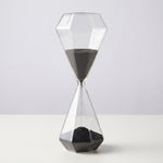 Cargar imagen en el visor de la Galería, Reloj de arena Tebas de vidrio de 30 minutos con arena negra cayendo sobre una mesa blanca.