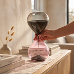 Cargar imagen en el visor de la Galería, Mano sosteniendo el Reloj de Arena Marea de Cristal Rosa Decorativo 27 cm con arena oscura en una mesa.