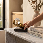 Cargar imagen en el visor de la Galería, Mano colocando la figura Saria, escultura decorativa de mano dorada con esfera sobre mesa de mármol.