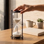 Cargar imagen en el visor de la Galería, Reloj de Arena Decorativo Atria en Cristal y Metal Negro con arena blanca sobre un escritorio de madera.