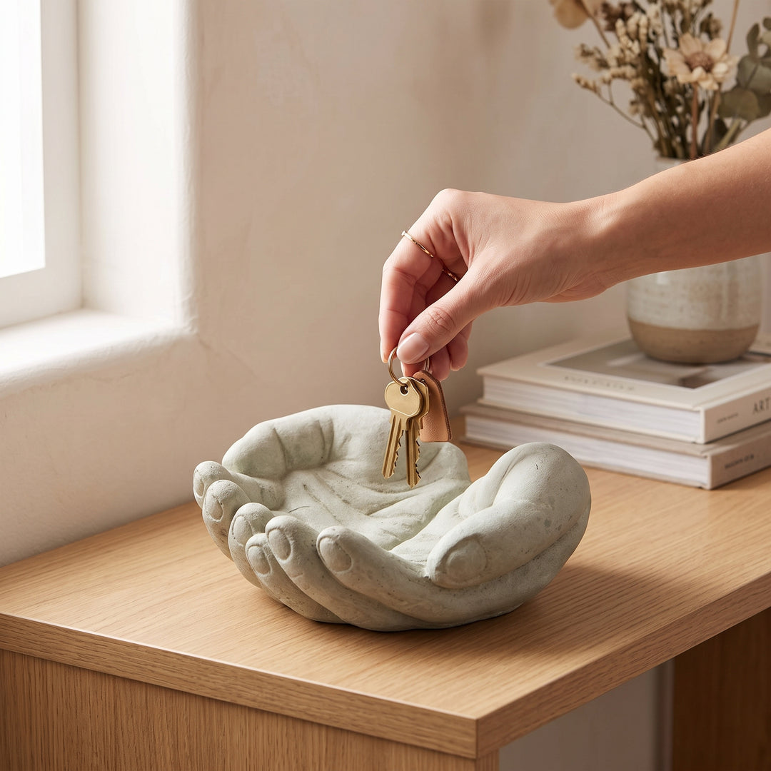 Mano colocando llaves en la escultura decorativa manos chicas color cemento sobre una mesa de madera.