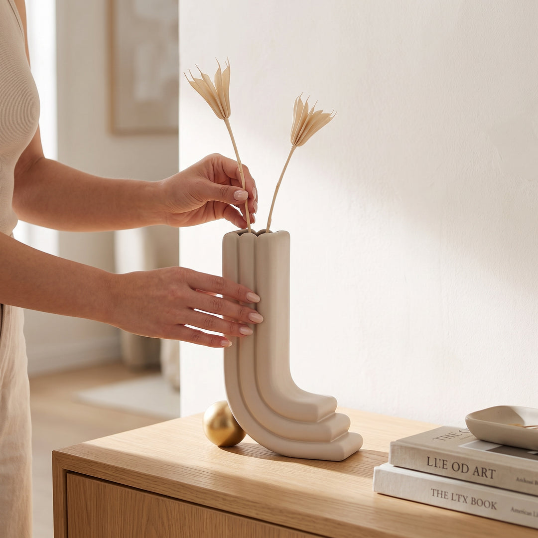 Manos colocando flores en el jarrón decorativo Oguz beige moderno sobre una mesa de recibidor.