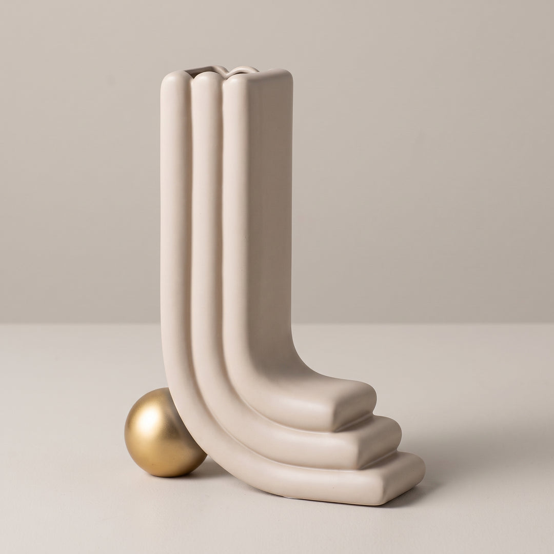 Jarrón decorativo Oguz beige moderno en forma de L con base de esfera dorada sobre fondo beige.