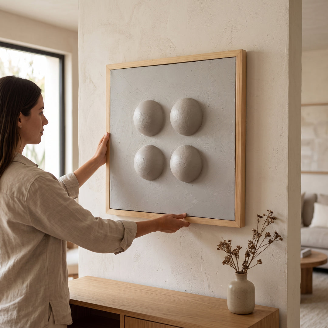 Mujer colocando cuadros perlas decorativos de 50x50 cm para sala y dormitorio en pared beige.