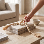 Cargar imagen en el visor de la Galería, Mano colocando la guirnalda de cuentas decorativas Mala Tashi de MDF 77cm sobre libros en una mesa de centro.