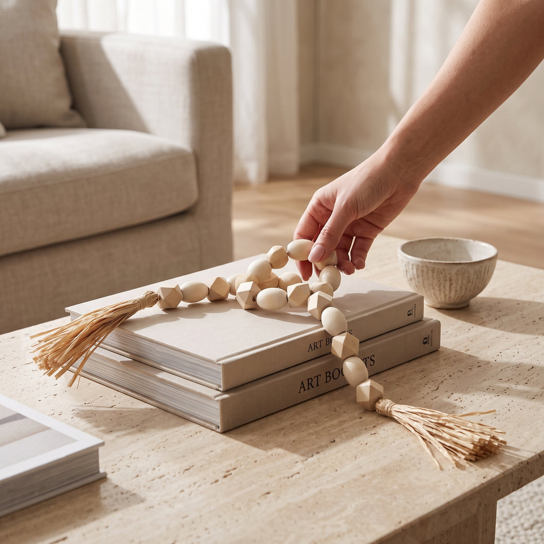 Mano colocando la guirnalda de cuentas decorativas Mala Tashi de MDF 77cm sobre libros en una mesa de centro.