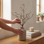Cargar imagen en el visor de la Galería, Manos colocando una rama decorativa artificial de magnolia en un florero de cerámica.