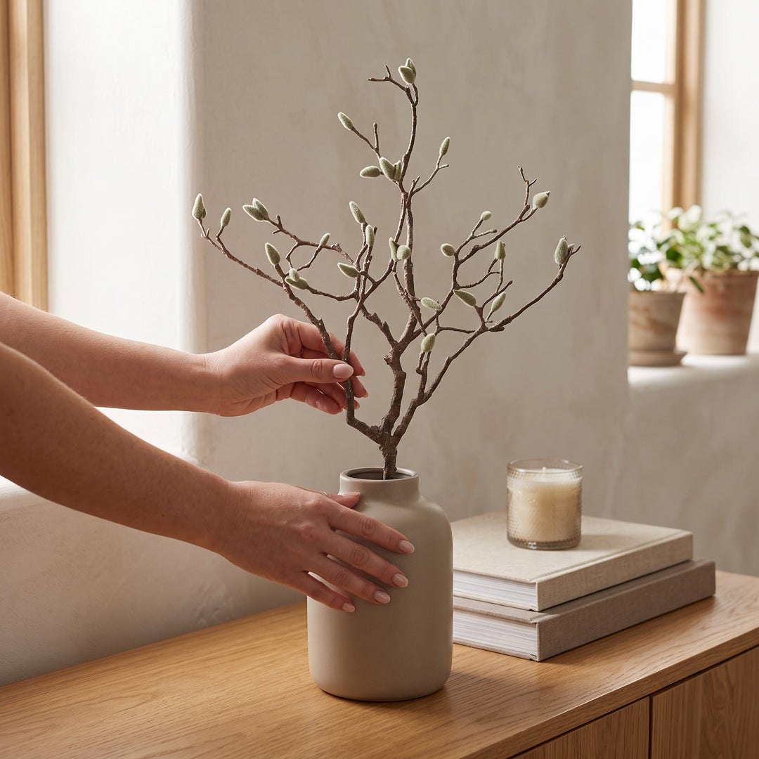 Manos colocando una rama decorativa artificial de magnolia en un florero de cerámica.