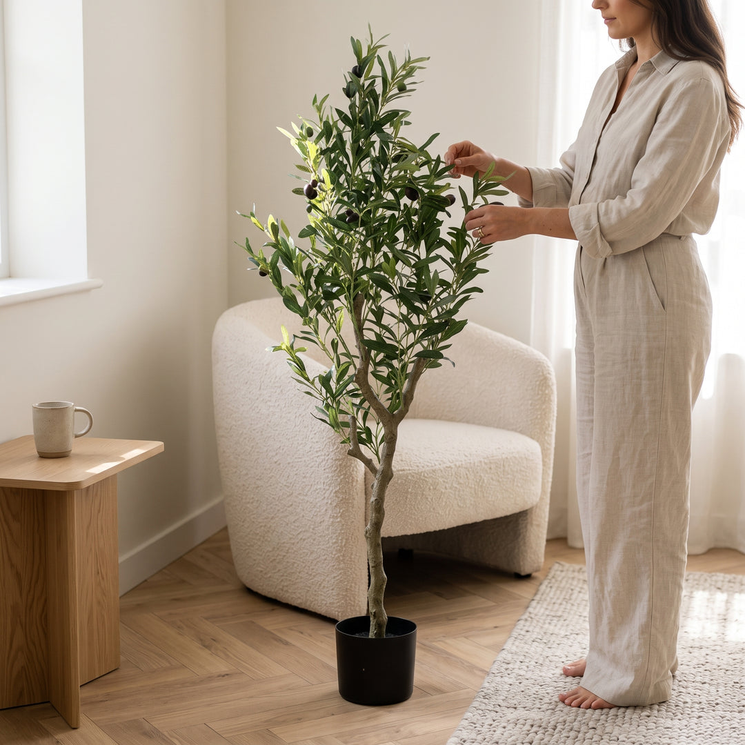 Mujer ajustando la planta decorativa artificial olivo mediano de 150cm en una sala luminosa.