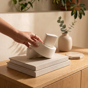 Mano colocando el accesorio decorativo de cerámica nudo blanco sobre libros en un mueble de madera.