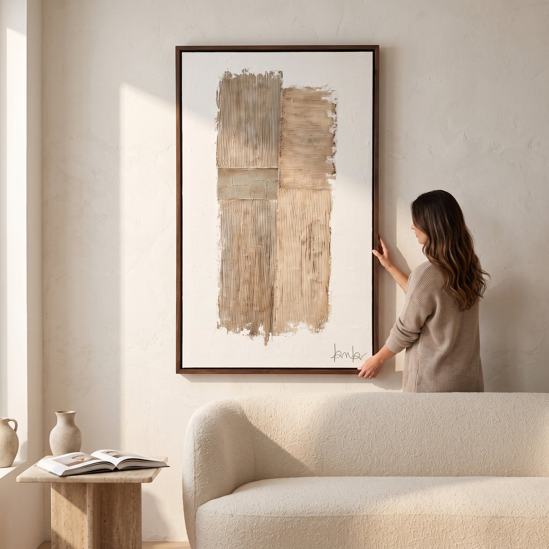 Mujer colgando el cuadro decorativo Kokay II de 0.90x1.50 cm sobre un sofá en un salón moderno.