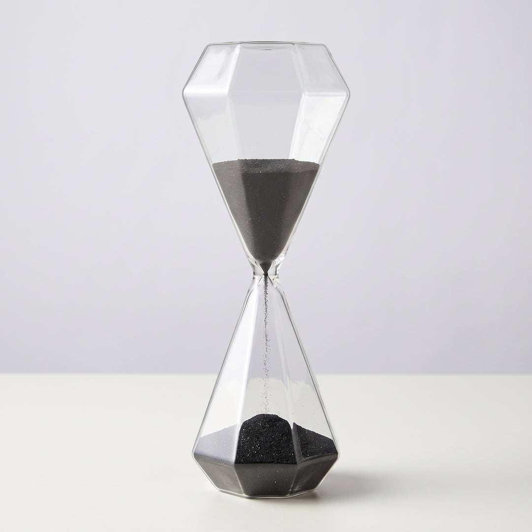 Reloj de arena Tebas de vidrio de 30 minutos con arena negra cayendo sobre una mesa blanca.