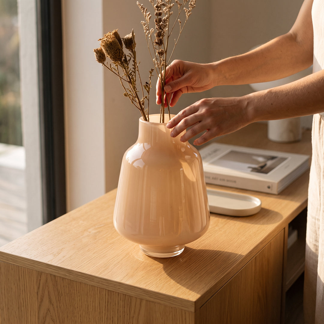 Manos colocando flores secas en el jarrón Luma Beige de vidrio decorativo sobre un mueble de madera.