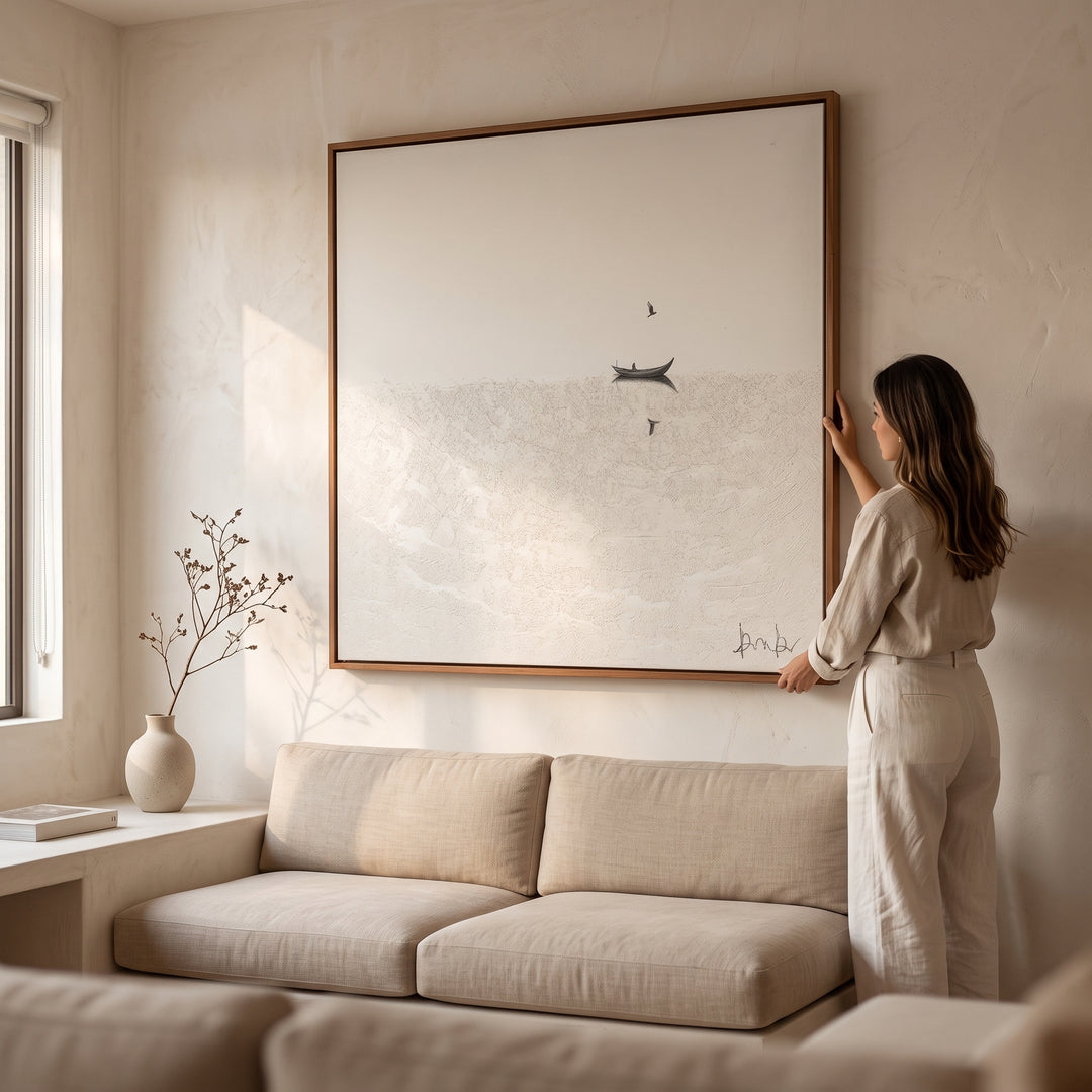 Mujer ajustando el cuadro decorativo A La Deriva con marco de madera sólida 140 X 140 sobre un sofá beige.