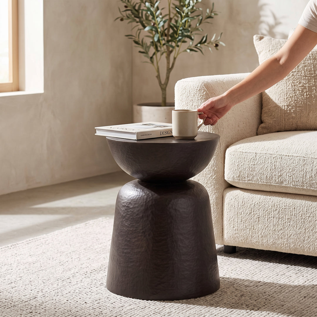 Mesa auxiliar Gladis compacta para sala o dormitorio en tono oscuro junto a un sillón beige con taza y libro.