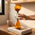 Cargar imagen en el visor de la Galería, Mano sosteniendo un reloj de arena decorativo de cristal de 35cm sobre libros en una mesa.