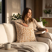 Cojín decorativo Moritz 50x50 cm con estampado simétrico beige en un sofá de sala junto a una mujer.