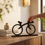 Cargar imagen en el visor de la Galería, Mano acomodando una bicicleta decorativa de mármol negro sobre un mueble de madera junto a libros de arquitectura.