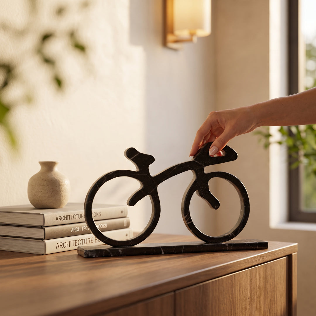 Mano acomodando una bicicleta decorativa de mármol negro sobre un mueble de madera junto a libros de arquitectura.