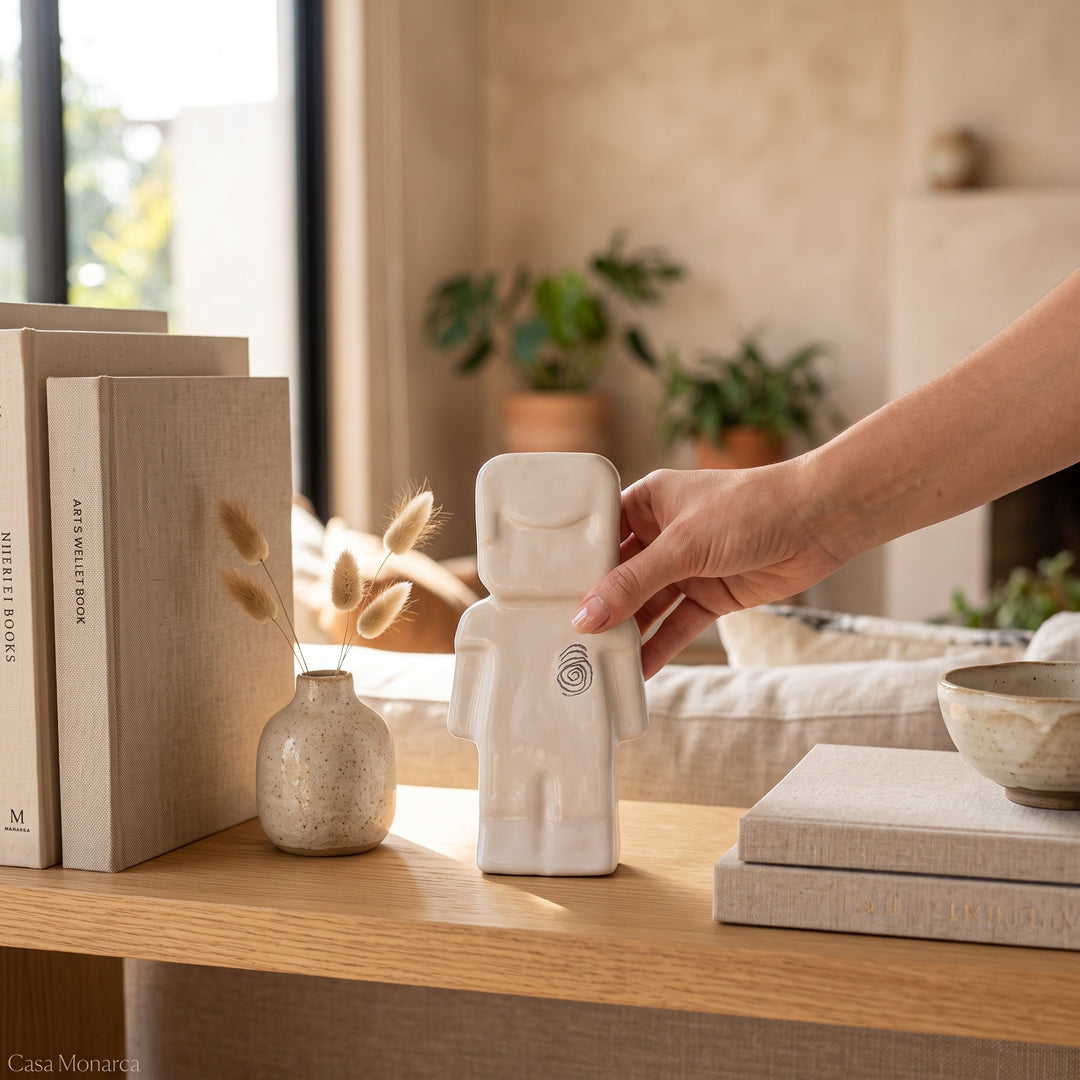 Mano colocando la escultura de cerámica blanca Huella de Sustento en una repisa de madera junto a libros.