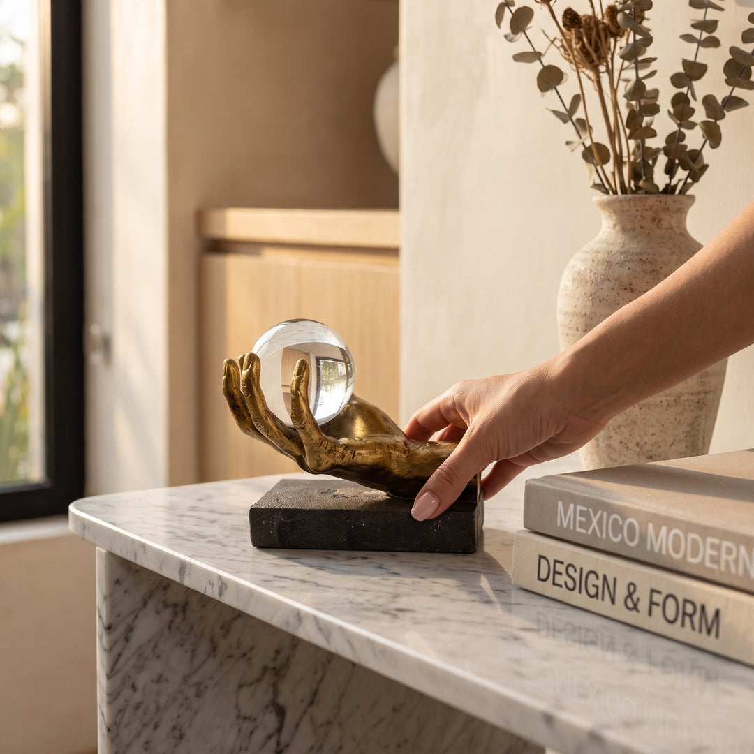 Mano colocando la figura Saria, escultura decorativa de mano dorada con esfera sobre mesa de mármol.