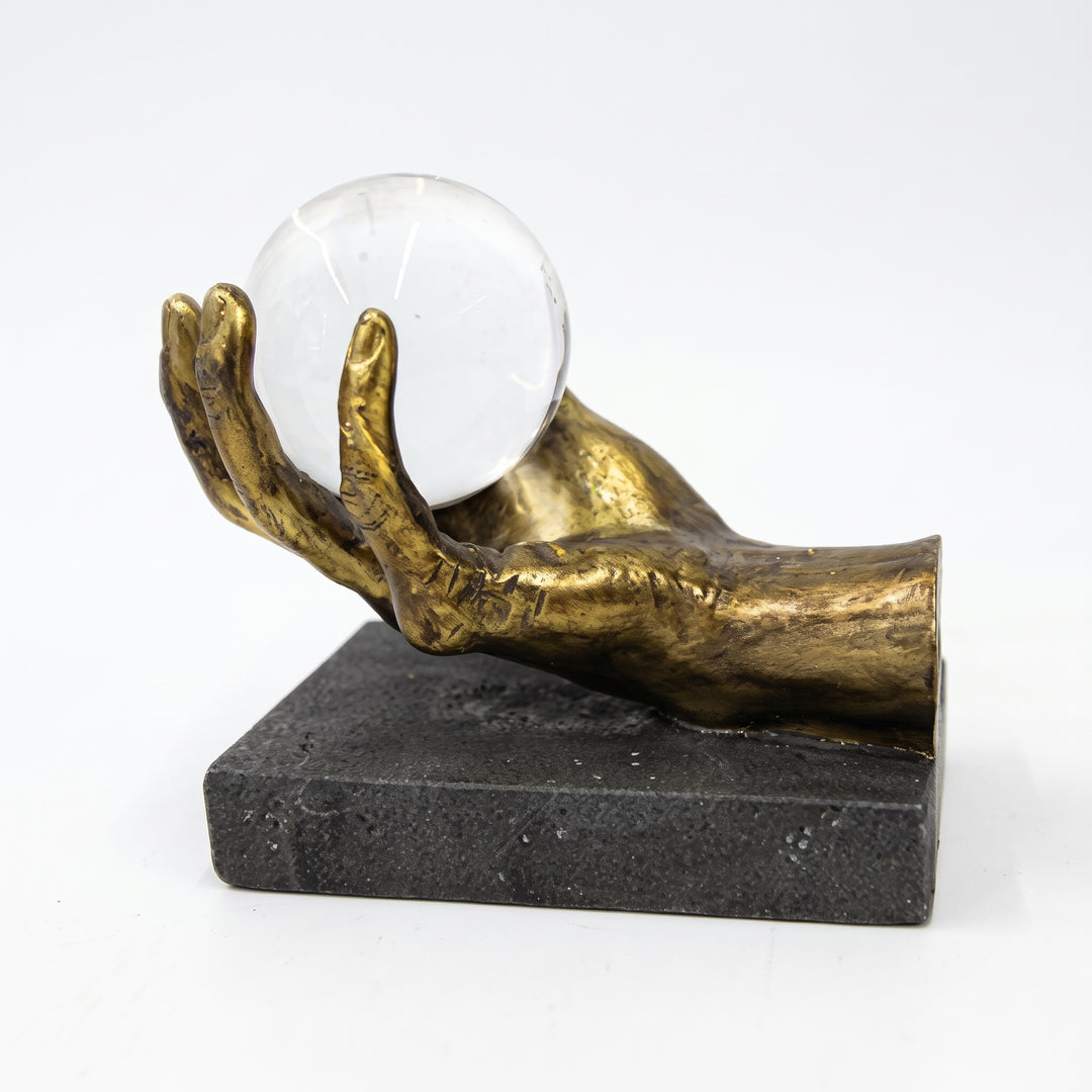 Figura Saria escultura decorativa de mano dorada con esfera de cristal sobre base oscura.