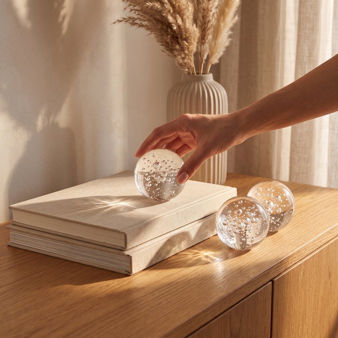 Mano colocando el juego de 3 esferas decorativas de cristal Liora para mesa sobre libros en un mueble de madera.