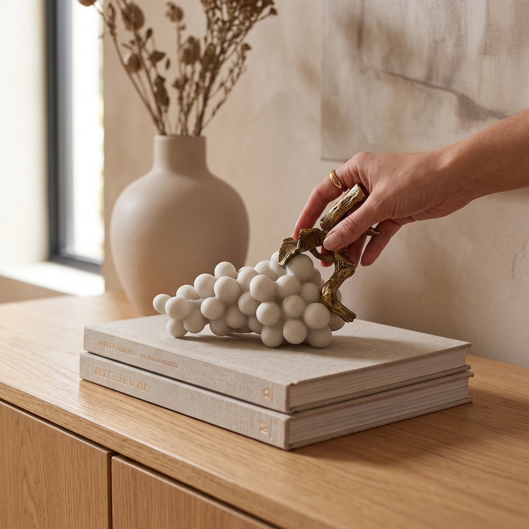 Figura Solen escultura uvas blanco y dorado de poliresina sobre libros decorativos en un mueble de madera.